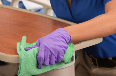 Gloved hands wiping down a hospital room table with a green cleaning cloth, providing healthcare support services.