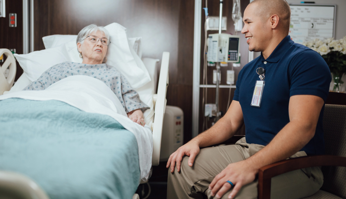 Elderly patient in hospital bed talking with smiling male healthcare worker in blue uniform.
