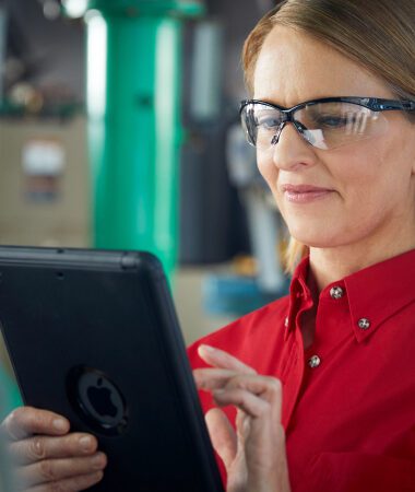 Focused woman in safety glasses using tablet in industrial workplace setting
