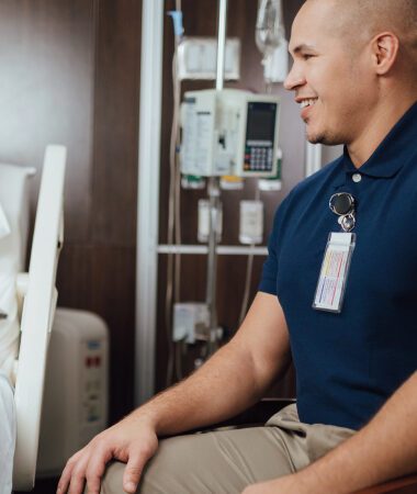 Smiling healthcare worker sitting in hospital room, wearing blue scrubs with ID badge attached