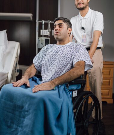 Patient in hospital gown and wheelchair attended by healthcare professional in medical setting