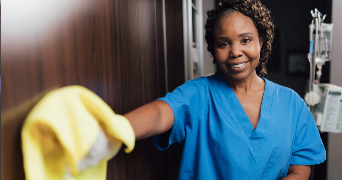 Smiling healthcare worker in blue scrubs wiping surface, promoting cleanliness in medical setting