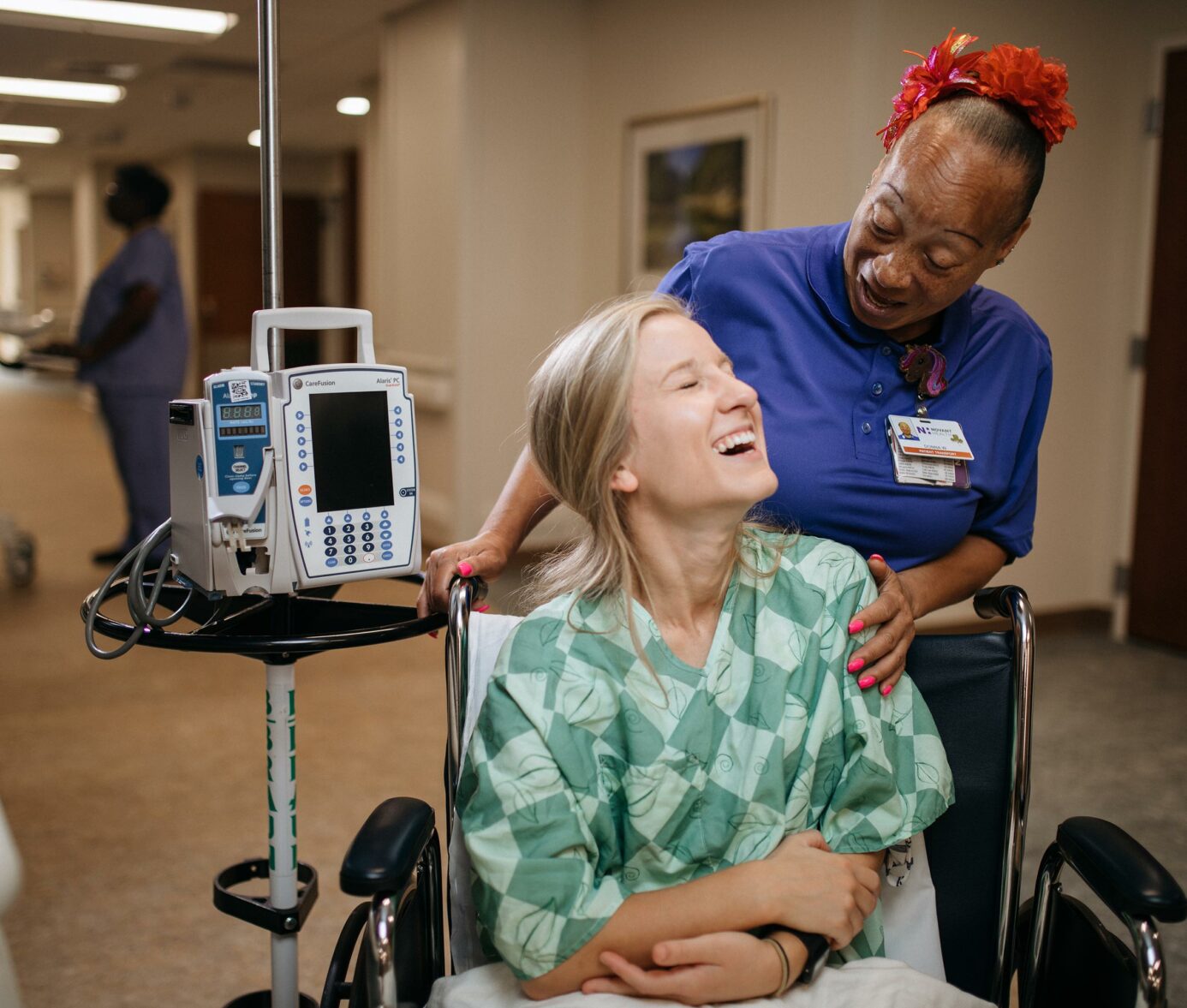 Blonde female patient in wheelchair laughing with African American female healthcare worker in hospital hallway.