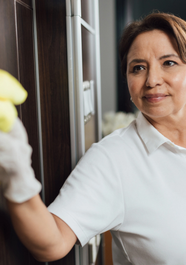 Smiling mature woman cleaning window in white uniform, representing healthcare support services.