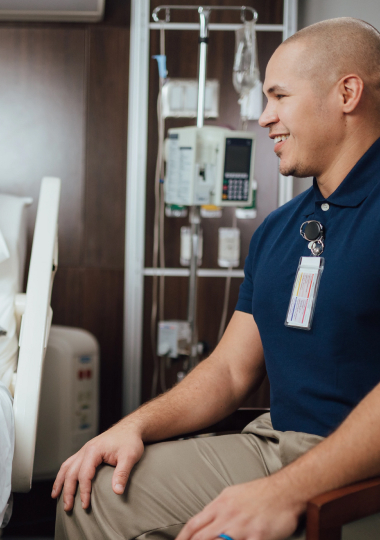 Smiling healthcare worker seated in patient's room, wearing ID badge and navy blue polo shirt.